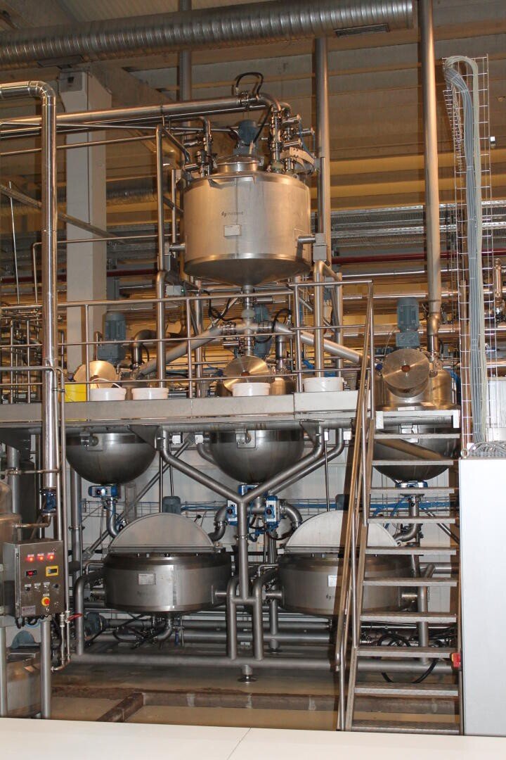 Large stainless steel industrial tanks and pipes in a food or chemical processing facility, with control panel, stairs, and multiple interconnected vessels and machinery.