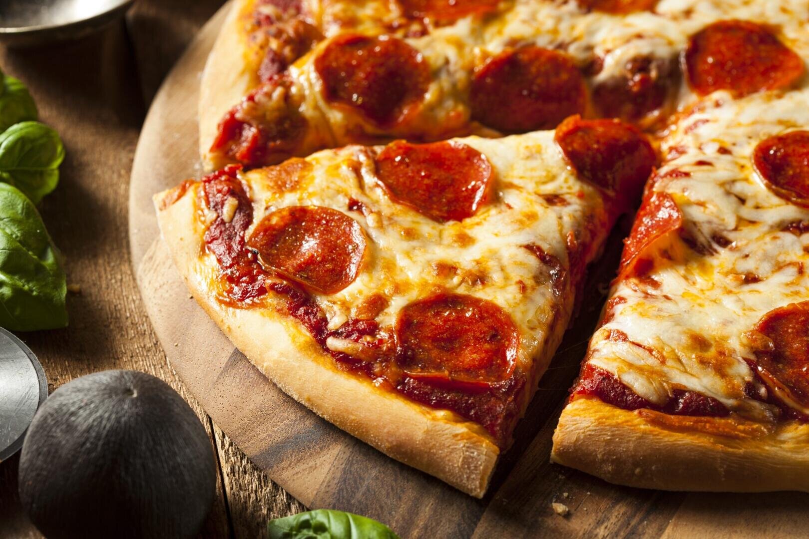 A close-up of a pepperoni pizza with melted cheese, sliced into pieces on a wooden board. The pizza sits on a rustic table with basil leaves and a black olive nearby.