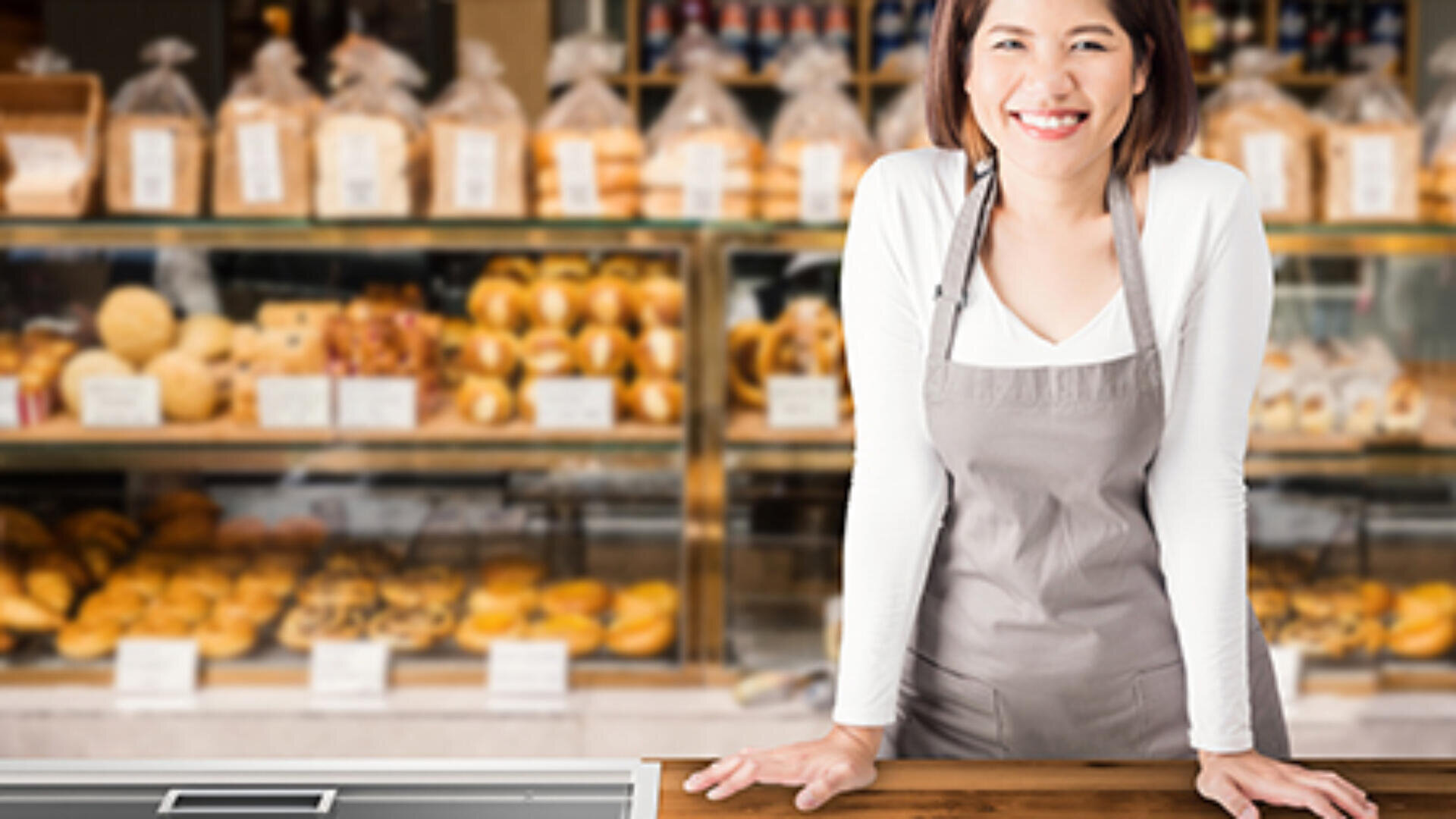 Eine lächelnde Frau mit einer grauen Schürze steht hinter einer Theke in einer Bäckerei, in der im Hintergrund Regale mit verschiedenen Brotsorten und Gebäck ausgestellt sind.