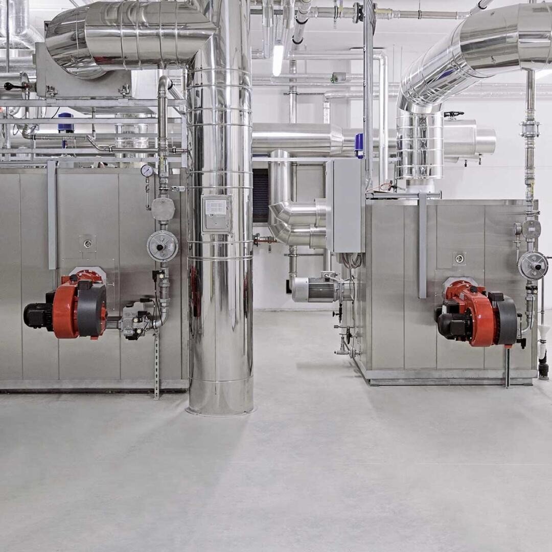 Ein sauberer, moderner industrieller Kesselraum mit großen Edelstahlrohren, Metalltanks und Maschinen. Zwei rote und schwarze Brennereinheiten sind an der Vorderseite von rechteckigen Kesseln auf beiden Seiten des Bildes angebracht.