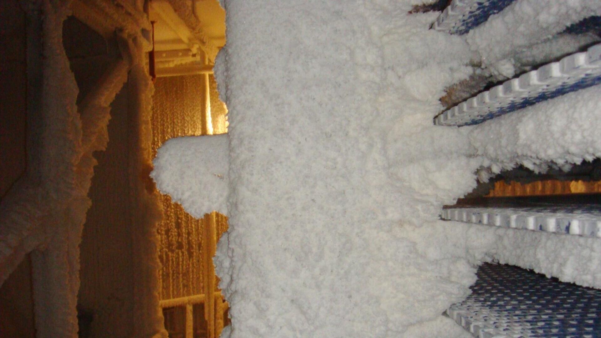 Eine dicke Frostschicht bedeckt Metallrohre und Maschinen in einer Industrieanlage. An den Wänden und Geräten bildet sich Eis, wodurch die gesamte Umgebung wie aus Schnee aussieht.