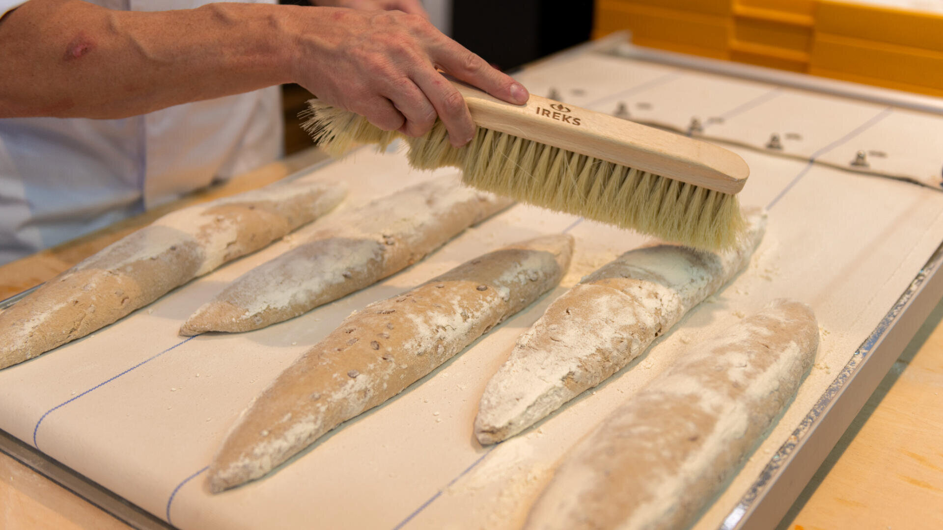 Eine Person stäubt mit einer großen Bürste Mehl von mehreren ungebackenen Baguettes, die in einer Bäckerei auf einem Tablett angeordnet sind.