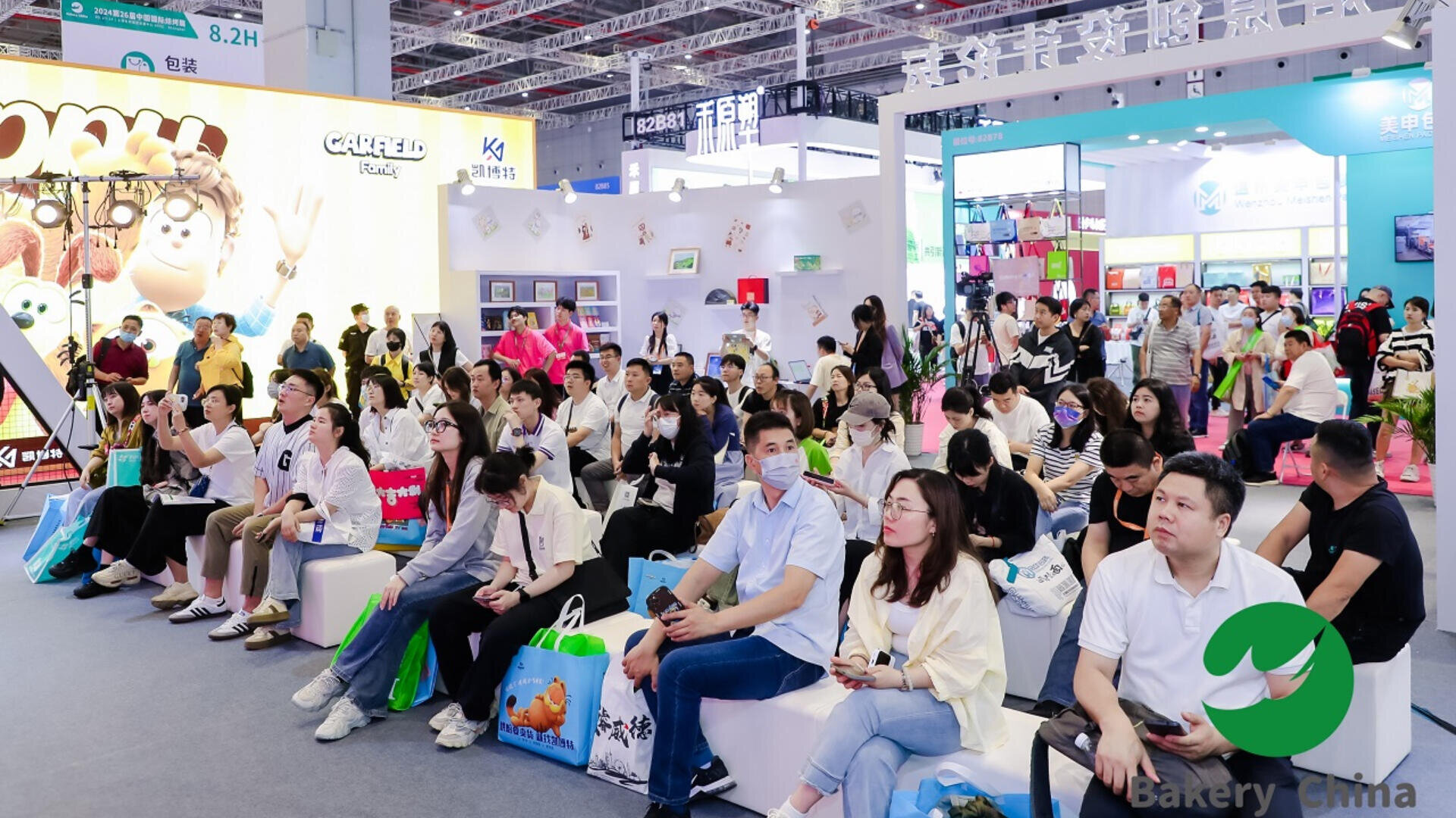 A large group of people sit and stand in a brightly lit exhibition hall, attentively watching a presentation. Booths with colorful displays and banners are visible in the background.