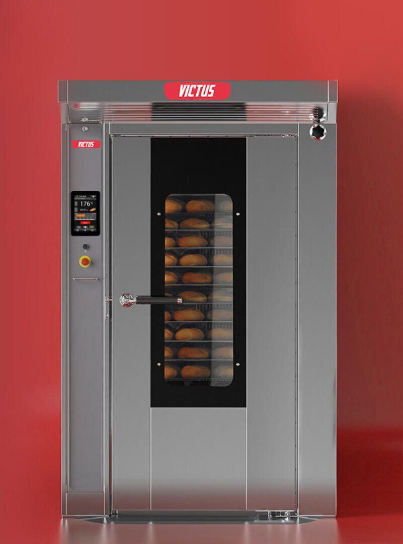 A large industrial stainless steel bread oven with a glass door showing multiple trays of loaves baking inside, set against a plain red background. The oven displays the brand VICTUS at the top.