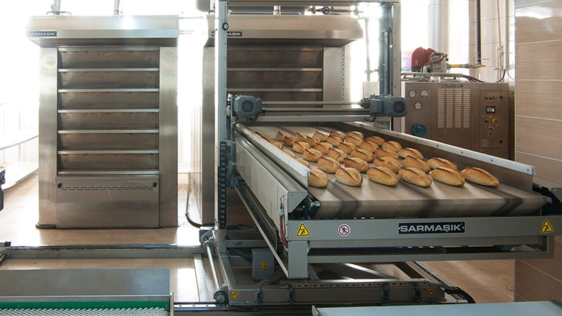 Industrial bread-making machine with loaves of bread on a conveyor belt, next to large ovens and control panels in a bakery production facility.