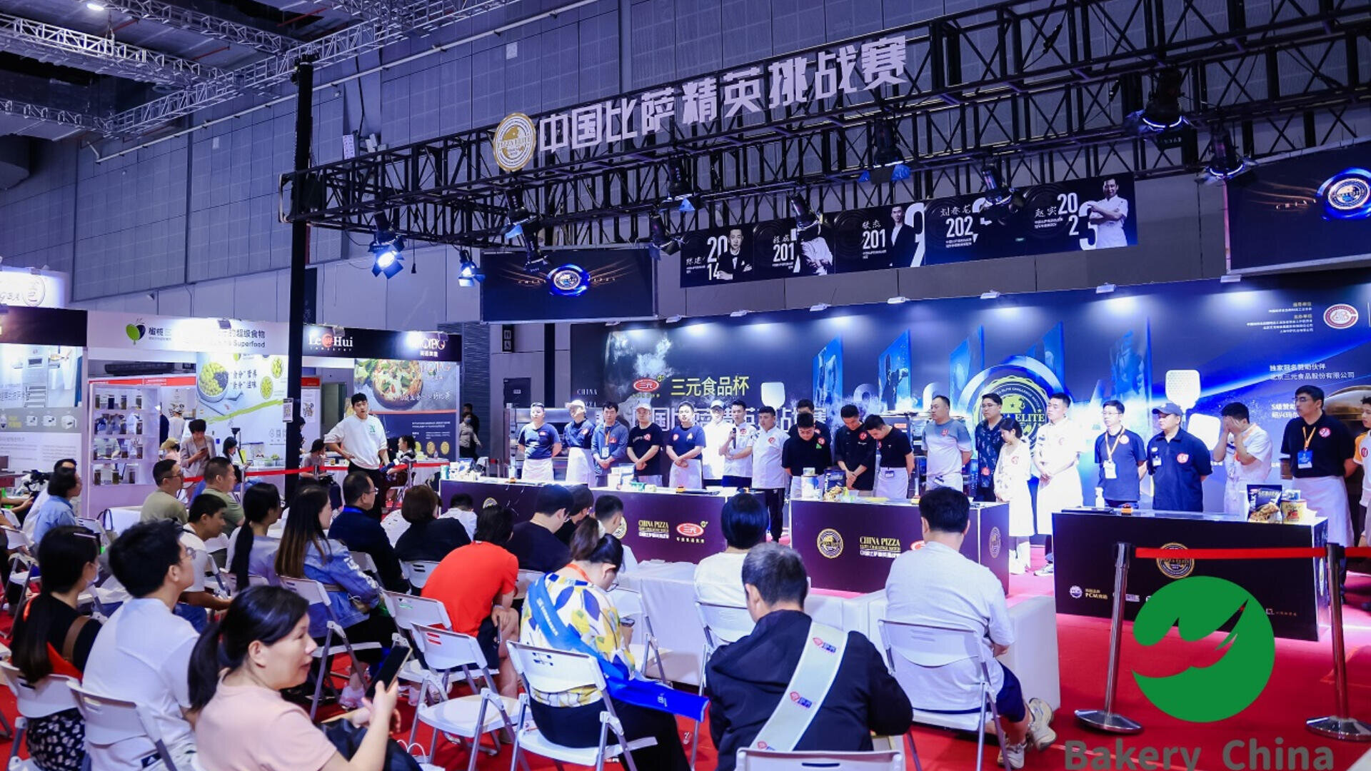 A large indoor event with many seated attendees watching a baking competition on stage. Contestants in white coats work at tables under bright lights, with banners and displays in the background.