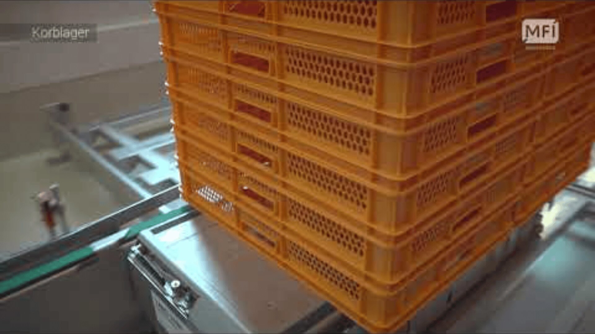 A stack of orange plastic crates sits on a metal conveyor system in an industrial setting. The crates have ventilation holes and the machinery appears to be part of an automated process.