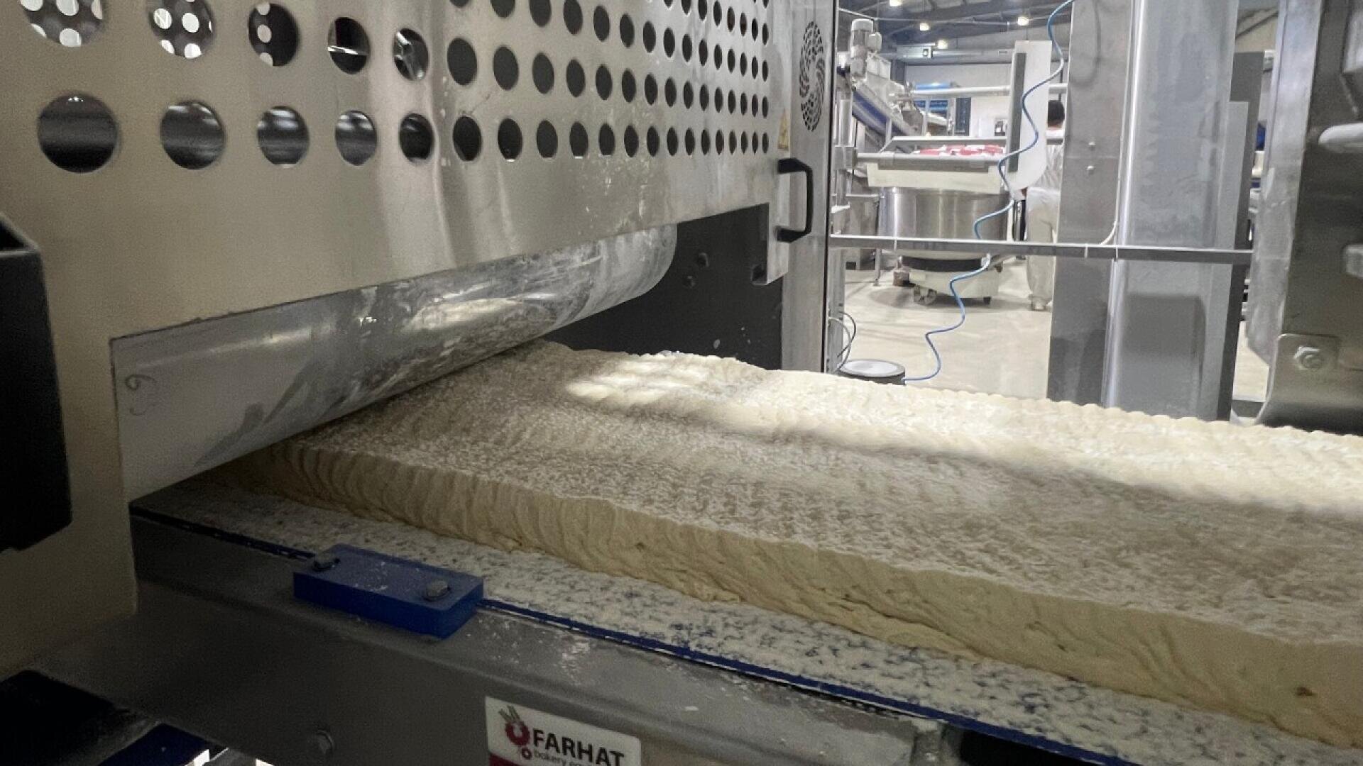 Ein großes Teigband läuft durch eine Industriemaschine in einer Fabrikumgebung, wobei im Hintergrund Metallflächen und -geräte zu sehen sind. Die Umgebung wirkt sauber und geordnet.