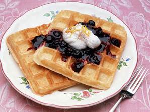 Zwei quadratische Waffeln mit Blaubeersoße und einem Klecks Schlagsahne werden auf einem blumengeschmückten Teller serviert, der auf einem rosa Tischtuch steht, mit einer Gabel neben dem Teller.