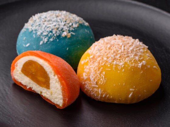 Three colorful mochi desserts—one blue, one yellow, and one orange with a mango-like filling—lie on a dark plate, each sprinkled with a pinch of coconut flakes.
