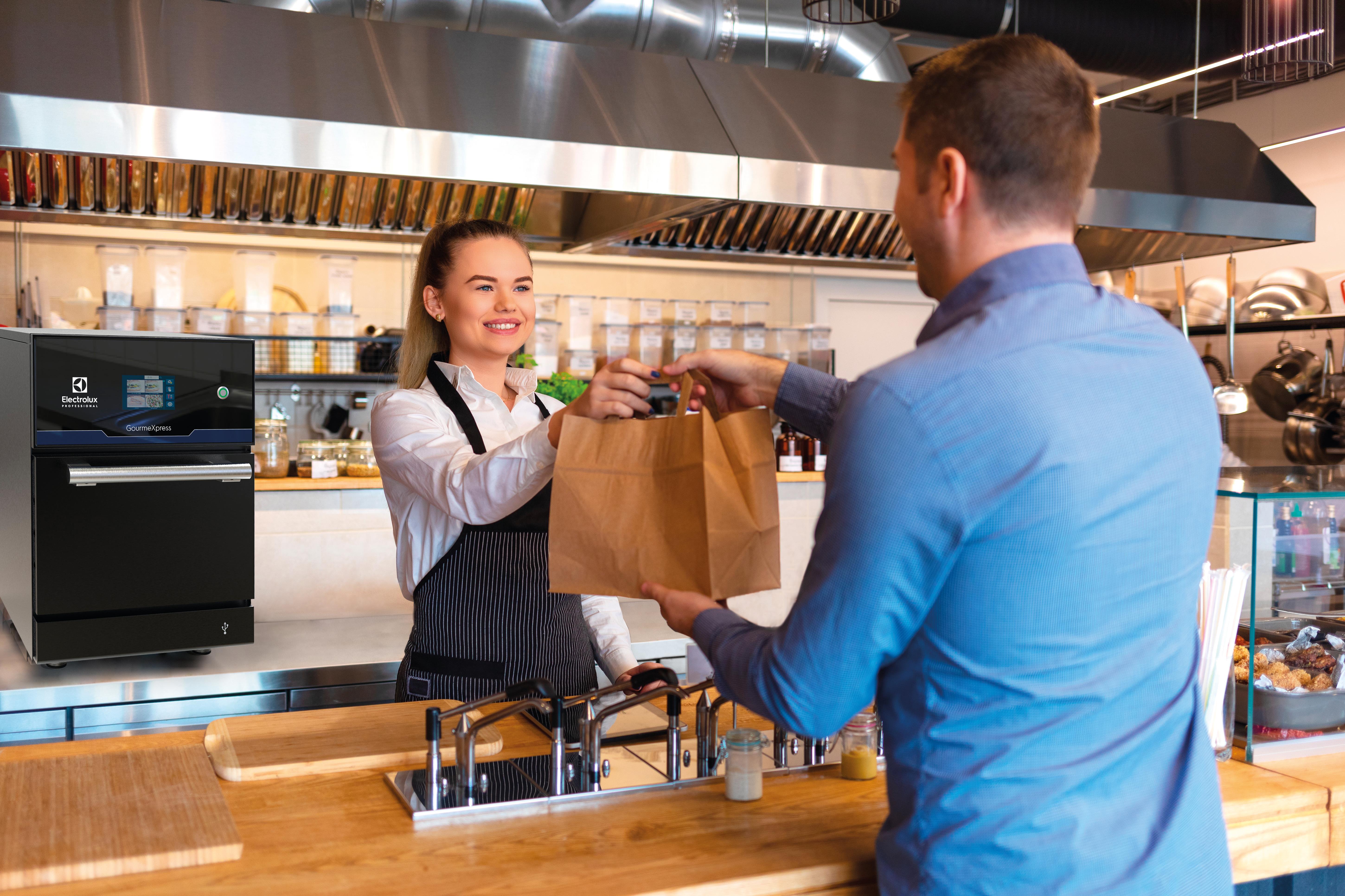 A smiling female employee hands a paper bag of food to a male customer across the counter in a modern restaurant kitchen. A black Electrolux appliance is visible on the counter to the left.