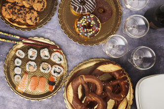 A table set with trays of assorted cookies, donuts, sushi, and large pretzels, alongside empty wine glasses and stacked white plates on a purple surface.