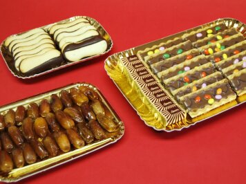 Three gold trays on a red surface: one with chocolate-dipped cookies, one with dates, and one with square biscuits topped with chocolate and colorful candies.