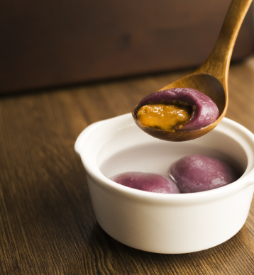 Auf einem Holzlöffel liegt ein violetter Klebreisball mit Füllung, über einer weißen Schüssel liegen auf einer Holzoberfläche zwei weitere Reisbällchen in einer klaren Flüssigkeit.