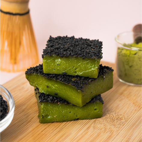 Drei quadratische Stücke grünen Matcha-Kuchens, garniert mit schwarzen Sesamkörnern, sind auf einem Holzbrett gestapelt, im Hintergrund stehen ein Bambusbesen und eine kleine Schale Matcha.