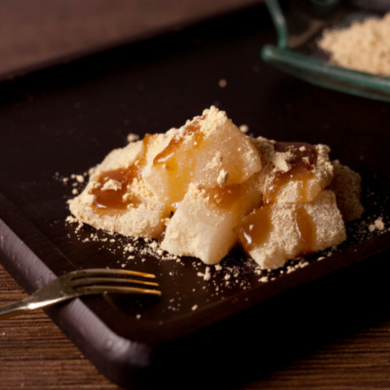 Drei Stücke durchsichtiges Mochi werden mit braunem Sirup und Kinako-Pulver bestreut und auf einem dunklen Holztablett serviert. Daneben liegt eine kleine Gabel. Im Hintergrund steht ein Schälchen mit zusätzlichem Pulver.