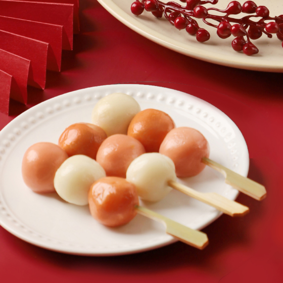 A white plate with three skewers of round dango balls, alternating between light brown and white, on a red surface with festive decorations in the background.