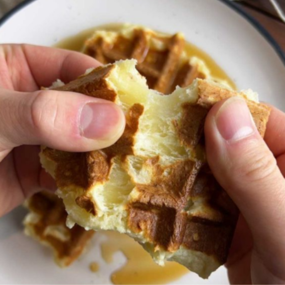 Zwei Hände zerbrechen eine Waffel über einem weißen Teller mit Sirup.