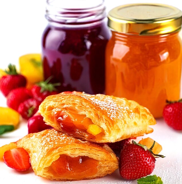 Two fruit-filled pastries are stacked in front of jars of red and orange jam, surrounded by fresh strawberries and some yellow fruit pieces on a white surface. The pastries are dusted with powdered sugar.