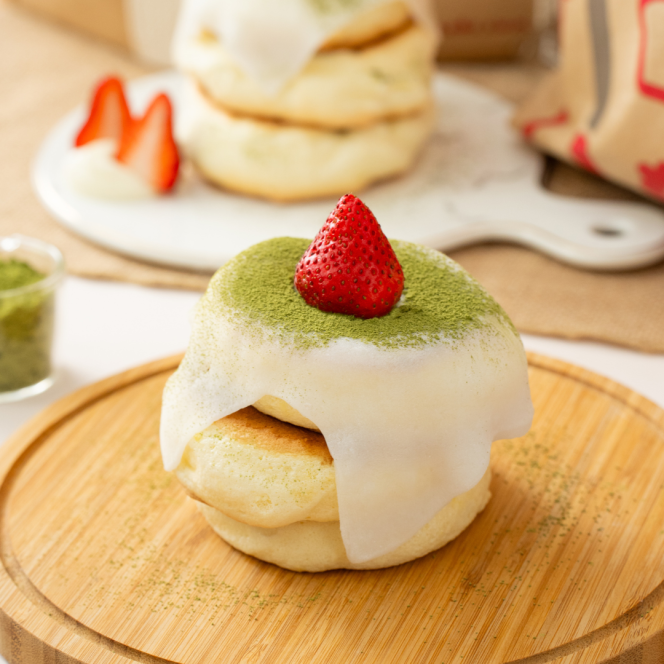 Ein fluffiger japanischer Pfannkuchen mit weißer Glasur, Matcha-Pulver und einer ganzen Erdbeere, serviert auf einem Holzteller mit weiteren Pfannkuchen und geschnittenen Erdbeeren im Hintergrund.