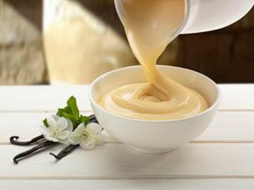 Ein cremiger, heller Vanillepudding wird aus einem Krug in eine weiße Schale auf einer weißen Holzfläche gegossen, neben der Vanilleschoten und weiße Blüten stehen.