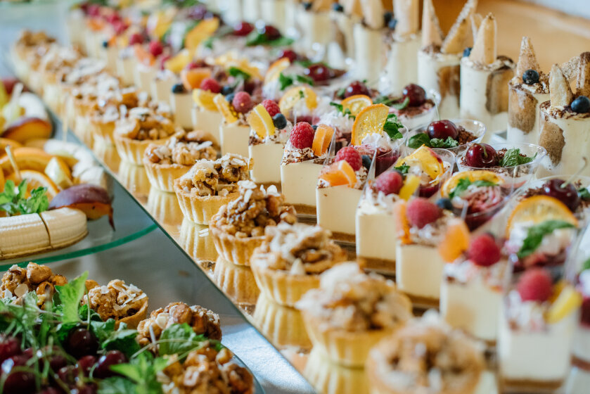 A colorful assortment of desserts, including tarts, cheesecakes, and pastries, topped with fresh fruits, nuts, and herbs, arranged on a buffet table.