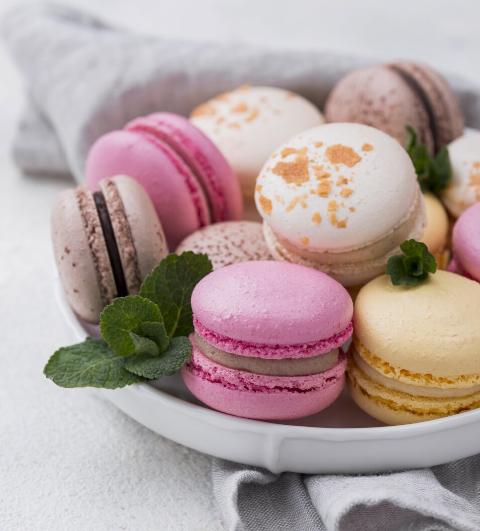 A white plate filled with colorful macarons in pink, yellow, white, and brown, garnished with fresh mint leaves, sits on a light gray cloth against a white background.