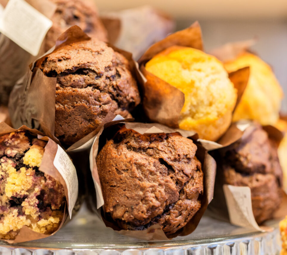 A close-up of assorted muffins, including chocolate and vanilla, wrapped in brown paper liners and arranged on a glass stand. The muffins appear fresh and moist, with textured, golden-brown tops.