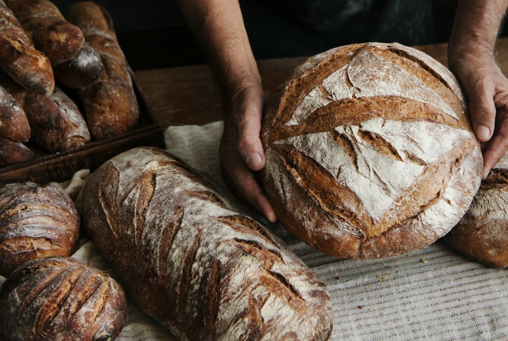 Eine Person hält einen runden Laib rustikalen Handwerksbrotes in der Hand, umgeben von anderen Broten auf einem mit Stoff bedeckten Tisch. Das Brot hat eine goldene Kruste mit Mehlbestäubung und dekorativen Rillen auf der Oberseite.