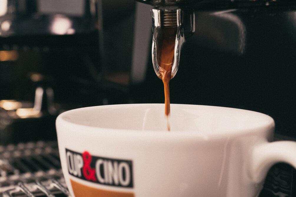 Nahaufnahme eines Espressos, der aus dem Ausguss einer Kaffeemaschine in eine weiße Tasse von CUP&CINO fließt. Dabei wird der kräftige, braune Kaffeestrahl während des Eingießens eingefangen.