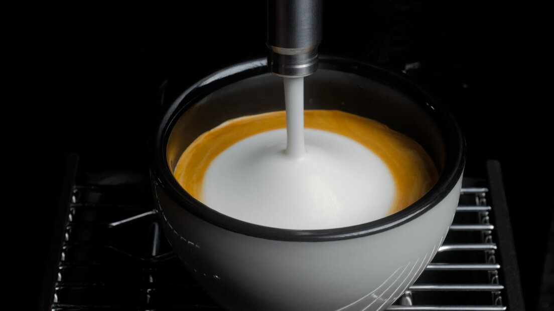 A close-up of a coffee cup being filled with steamed milk, creating a layer of foam on top of an espresso shot, sitting on a metal grate.