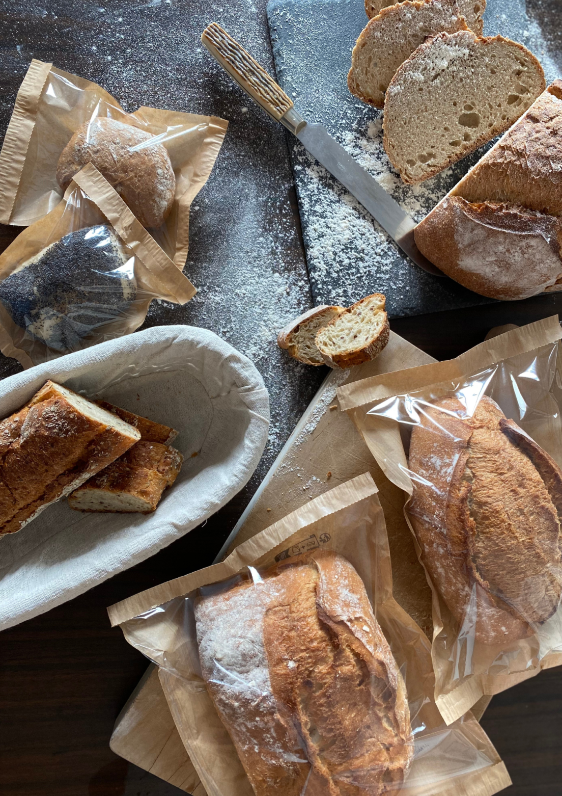 An assortment of freshly baked bread, some sliced and some whole, is displayed on a table. Several loaves are in paper bags, and a bread knife rests nearby with flour scattered on a cutting board.