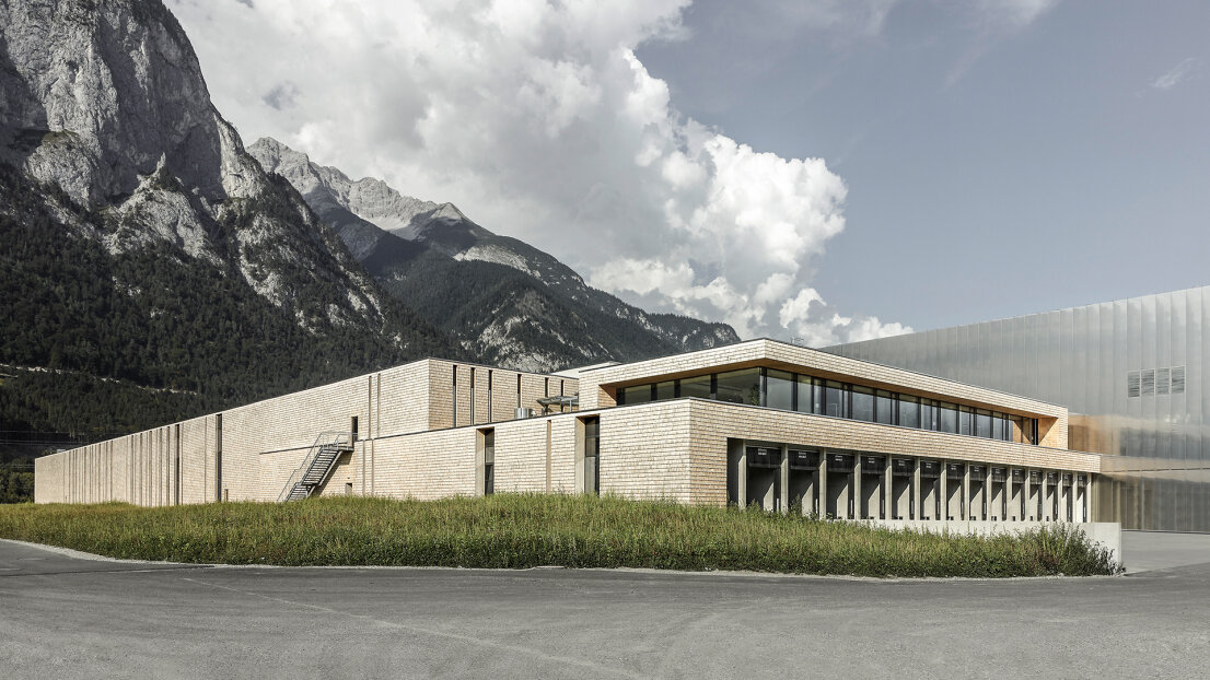 A modern, rectangular building with light-colored brick walls and large windows sits on a grassy plot, with tall mountains and a partially cloudy sky in the background.