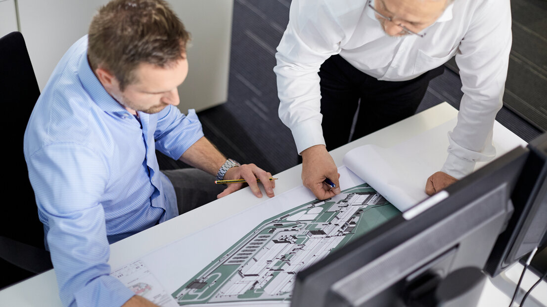 Two men in business attire are reviewing architectural plans at a desk. One is sitting, the other is standing, both concentrating on the drawings in front of a computer monitor.