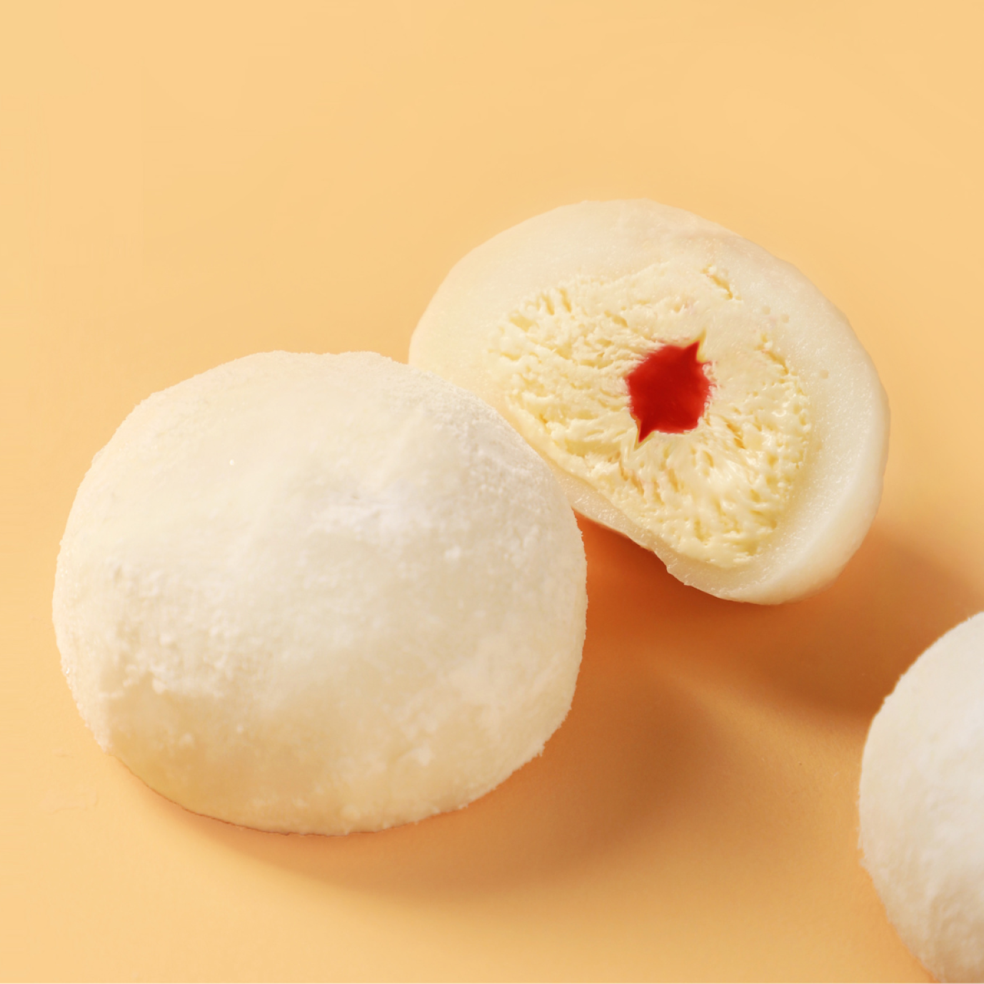 Zwei Mochi-Eiskugeln auf einem hellen Pfirsichhintergrund, eine ganz und eine aufgeschnitten, um cremiges Vanilleeis mit einer Füllung aus roten Früchten freizugeben.
