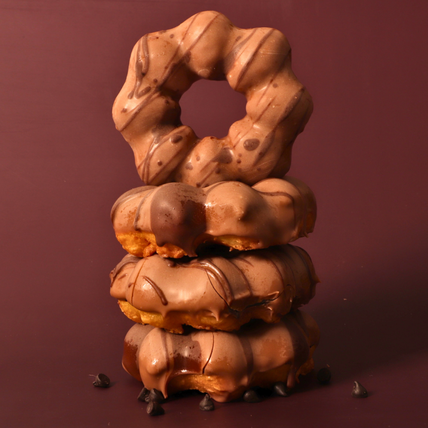 Three chocolate-glazed donuts are stacked on top of each other, with a fourth donut standing upright on top. Chocolate chips are scattered at the base, all set against a solid brown background.
