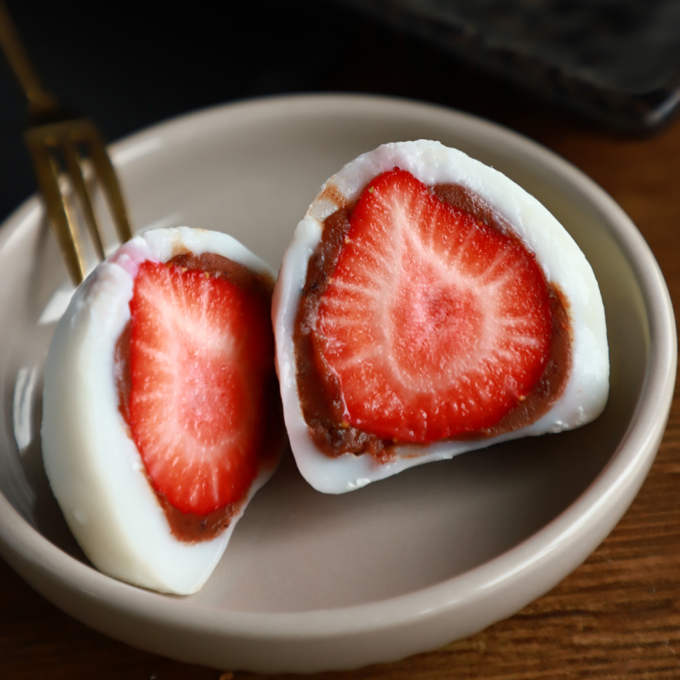 Eine Nahaufnahme von zwei Hälften Erdbeer-Daifuku – japanisches Mochi gefüllt mit einer ganzen Erdbeere und roter Bohnenpaste – auf einem kleinen beigen Teller mit einer Gabel im Hintergrund.