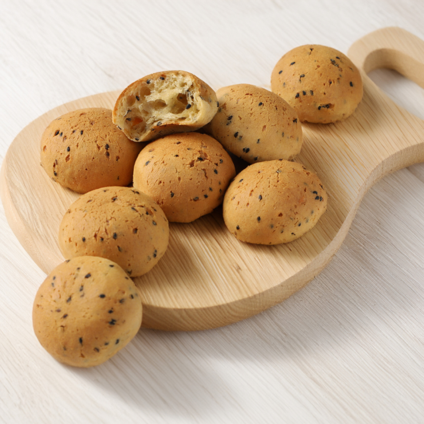 Runde, goldbraun gebackene Brötchen mit schwarzem Sesam auf einem hölzernen Servierbrett. Ein Brötchen ist aufgebrochen und gibt den Blick auf das weiche, luftige Innere frei. Das Ganze liegt auf einer hellen Unterlage.